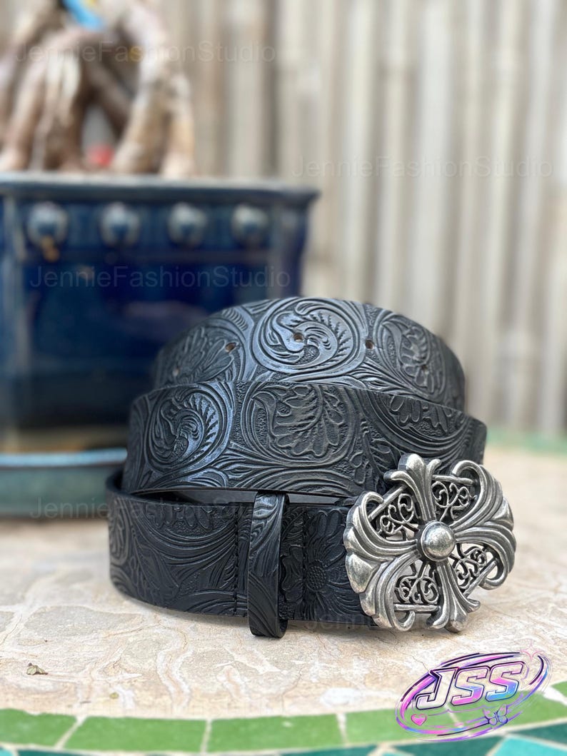 May include: Black leather belt with floral embossed pattern and a silver-colored buckle. The belt is rolled up, showcasing the intricate design. The buckle has a cross-like design.