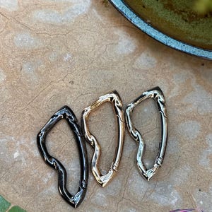 May include: Three metal carabiner clips in black, gold, and silver. The clips have an organic, flowing shape and are displayed on a textured, neutral-toned surface. The carabiners are likely used for attaching items.