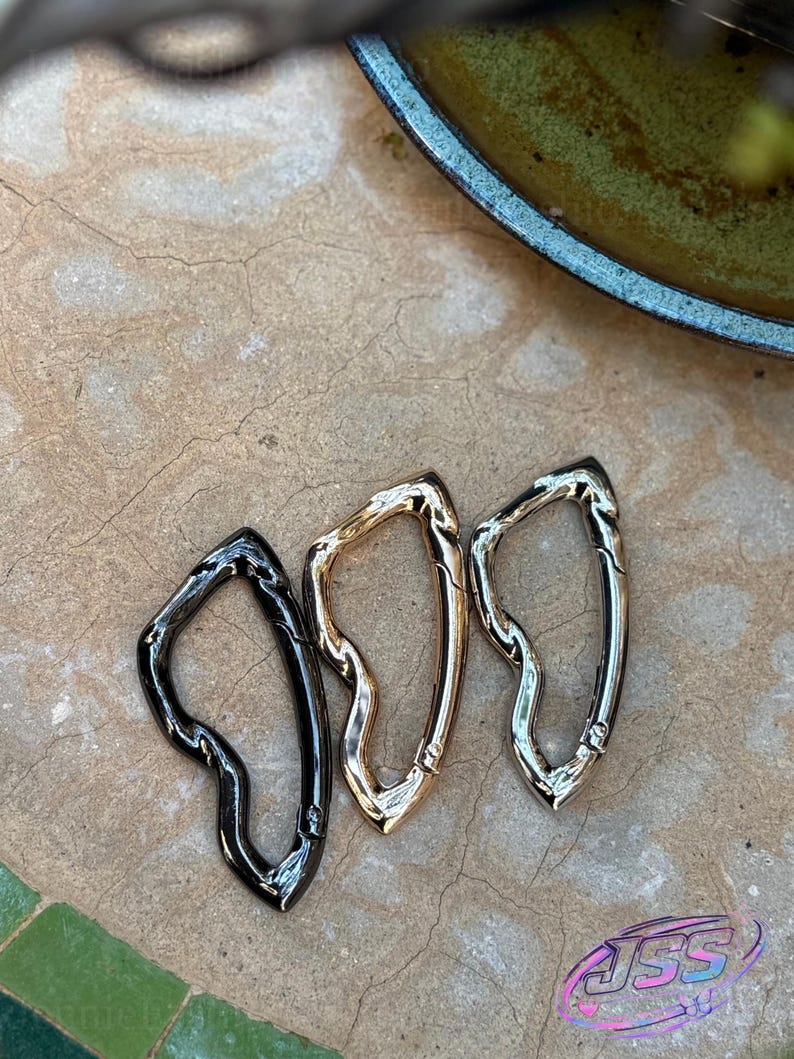 May include: Three metal carabiner clips in black, gold, and silver. The clips have an organic, flowing shape and are displayed on a textured, neutral-toned surface. The carabiners are likely used for attaching items.