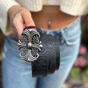 May include: Black leather belt with a silver-colored ornate buckle. The belt features a detailed floral pattern. The buckle has a cross-like design with a central stud. A stylish accessory for any outfit.