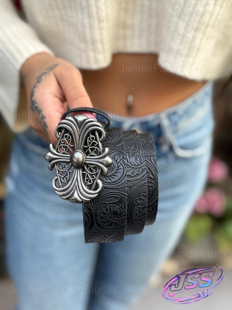 May include: Black leather belt with a silver-colored ornate buckle. The belt features a detailed floral pattern. The buckle has a cross-like design with a central stud. A stylish accessory for any outfit.