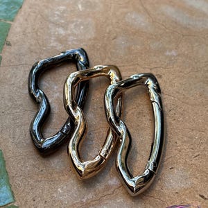 May include: Three metal swivel clasps in varying finishes: dark gunmetal, gold, and silver. The clasps have a unique, wavy design and are arranged on a textured, neutral-toned surface. The clasps are likely for use in bags or other accessories.