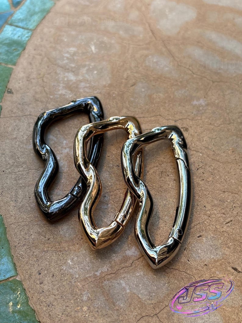 May include: Three metal swivel clasps in varying finishes: dark gunmetal, gold, and silver. The clasps have a unique, wavy design and are arranged on a textured, neutral-toned surface. The clasps are likely for use in bags or other accessories.