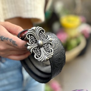 May include: Black leather belt with a large, ornate silver-colored cross buckle. The belt has a textured pattern and is held by a person. The buckle features intricate scrollwork and a central stud.