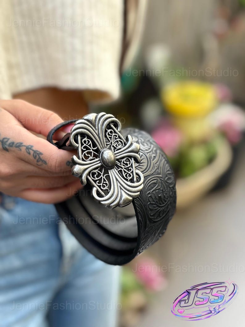 May include: Black leather belt with a large, ornate silver-colored cross buckle. The belt has a textured pattern and is held by a person. The buckle features intricate scrollwork and a central stud.