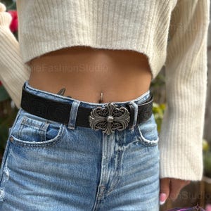 May include: Black leather belt with a silver-colored ornate buckle. The belt is worn with blue jeans and a cream-colored sweater. The buckle has a cross-like design.