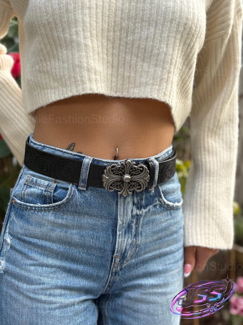 May include: Black leather belt with a silver-colored ornate buckle. The belt is worn with blue jeans and a cream-colored sweater. The buckle has a cross-like design.
