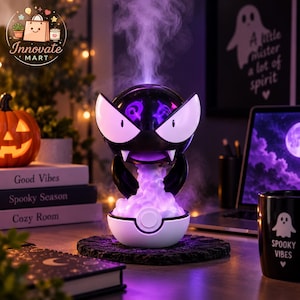 May include: A black and white spherical diffuser shaped like a cartoon creature with large white eyes and fangs, resting on a white base. A stream of purple-tinted mist rises from the top, while the base glows with a soft purple light.