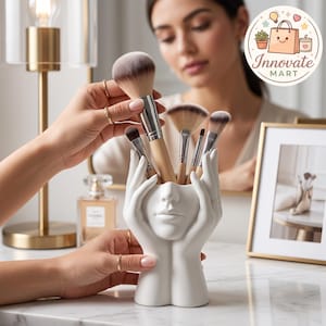 Puede incluir: Un soporte de cerámica blanca para brochas de maquillaje con forma de dos manos que sostienen un rostro. El soporte contiene varias brochas con mangos de madera y cerdas suaves. El diseño presenta un acabado mate liso y una forma escultural.
