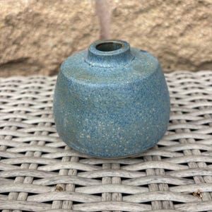 May include: A small, round ceramic vase with a speckled blue glaze. The vase features a wide, bulbous body that tapers towards a narrow, circular opening at the top. The surface texture appears matte with subtle variations in the blue and grey tones.