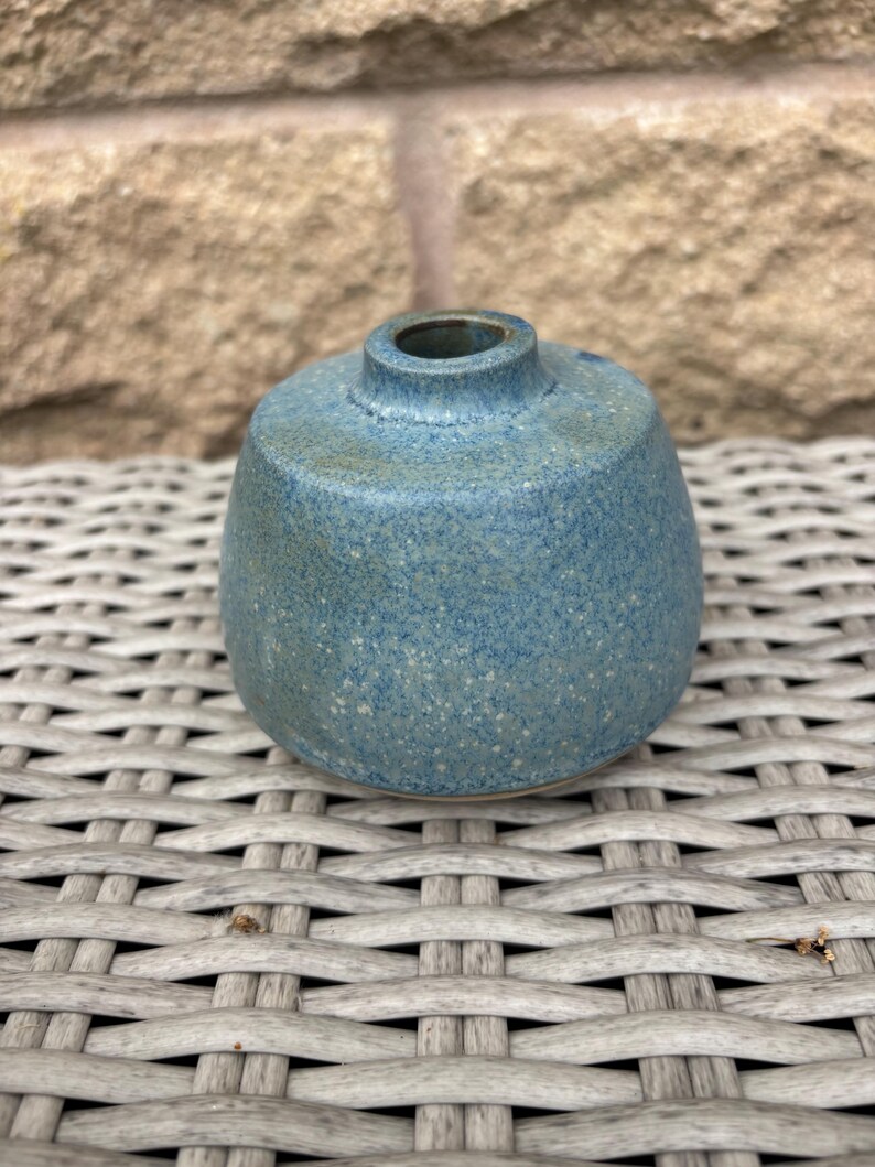May include: A small, round ceramic vase with a speckled blue glaze. The vase features a wide, bulbous body that tapers towards a narrow, circular opening at the top. The surface texture appears matte with subtle variations in the blue and grey tones.