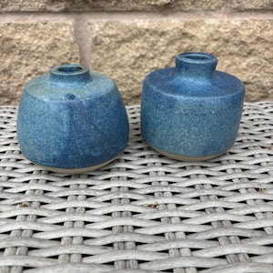 May include: Two small, round ceramic vases with a speckled blue glaze. Each vase features a short, narrow neck and a wide, rounded body. The base of each piece is unglazed, revealing a light tan colour. The surface texture appears slightly matte.