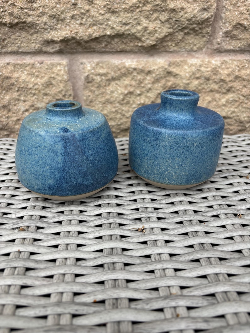 May include: Two small, round ceramic vases with a speckled blue glaze. Each vase features a short, narrow neck and a wide, rounded body. The base of each piece is unglazed, revealing a light tan colour. The surface texture appears slightly matte.