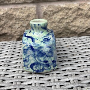 May include: A small, square-shaped ceramic vase with a narrow neck and opening. The piece features a glossy glaze with a light mint green base colour and dark blue, swirling, abstract brushstroke patterns covering the exterior surfaces.