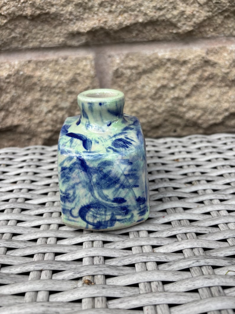 May include: A small, square-shaped ceramic vase with a narrow neck and opening. The piece features a glossy glaze with a light mint green base colour and dark blue, swirling, abstract brushstroke patterns covering the exterior surfaces.