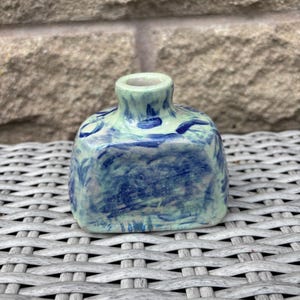 May include: A small, square-shaped ceramic vase with a short, narrow neck. The piece features a pale mint green base glaze with an abstract, hand-painted pattern of dark blue brushstrokes covering the front and sides of the vessel.