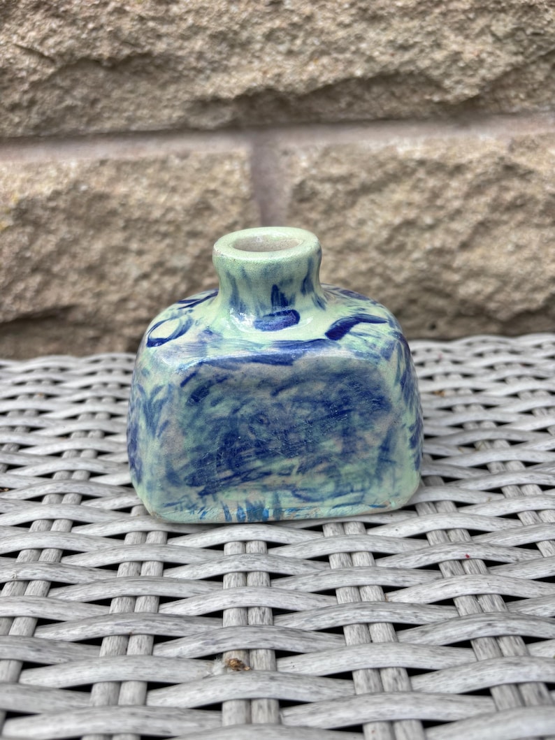 May include: A small, square-shaped ceramic vase with a short, narrow neck. The piece features a pale mint green base glaze with an abstract, hand-painted pattern of dark blue brushstrokes covering the front and sides of the vessel.