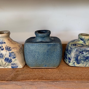 May include: Three small ceramic vases with square bases and rounded necks. The left vase is cream with blue floral patterns, the centre vase is solid textured blue, and the right vase features a marbled blue and cream glaze finish.