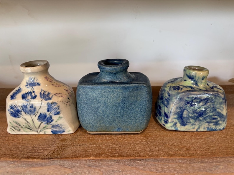 May include: Three small ceramic vases with square bases and rounded necks. The left vase is cream with blue floral patterns, the centre vase is solid textured blue, and the right vase features a marbled blue and cream glaze finish.