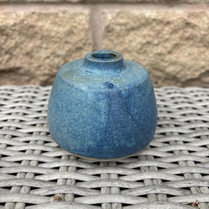May include: A small, round ceramic bud vase with a speckled blue glaze. The vase features a wide, bulbous body that tapers towards a narrow, circular opening at the top. The surface texture appears slightly uneven with variations in blue tones.