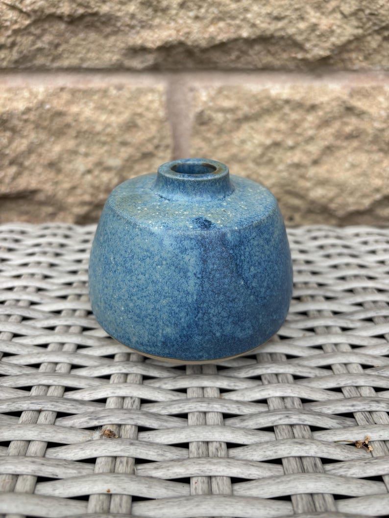 May include: A small, round ceramic bud vase with a speckled blue glaze. The vase features a wide, bulbous body that tapers towards a narrow, circular opening at the top. The surface texture appears slightly uneven with variations in blue tones.