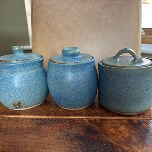 Puede incluir: Tres tarros de cerámica con tapa y esmalte azul moteado. Los dos tarros de la izquierda tienen cuerpos redondeados y pequeños pomos en las tapas, mientras que el tarro de la derecha es cilíndrico y tiene un asa arqueada en la tapa.