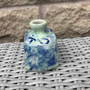 May include: A small, square-shaped ceramic vase with a short, narrow neck. The surface features a light green base colour with a mottled, hand-painted pattern of dark blue brushstrokes and abstract markings covering the exterior sides.