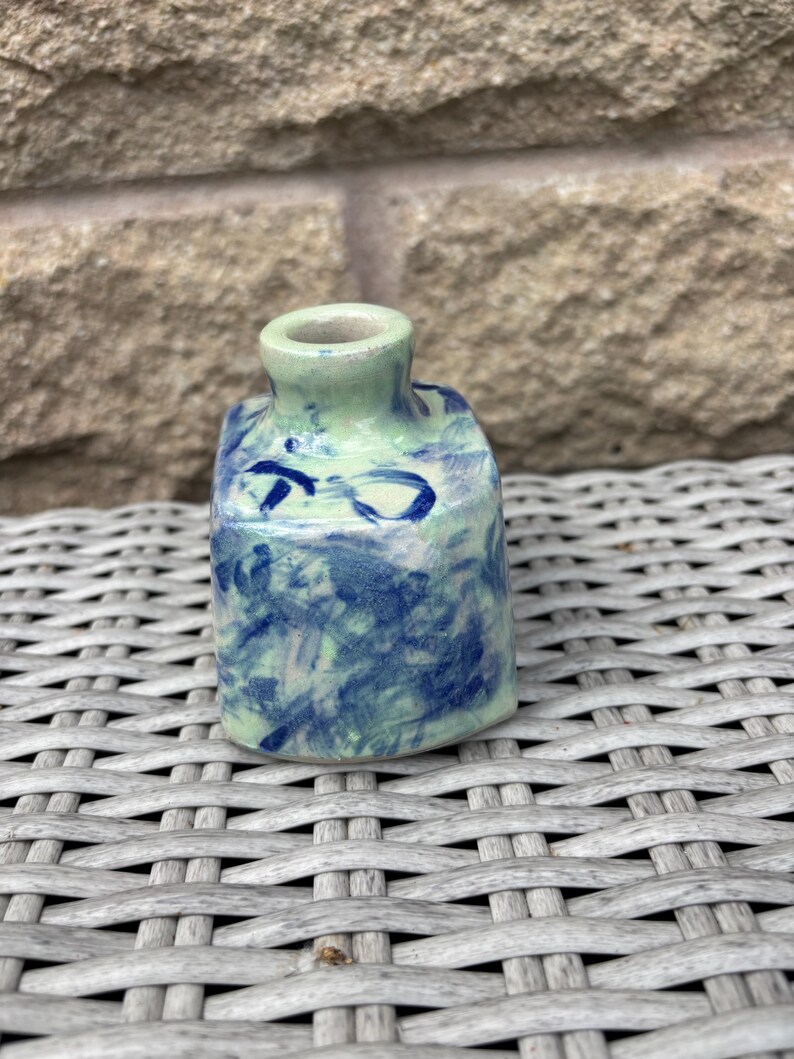 May include: A small, square-shaped ceramic vase with a short, narrow neck. The surface features a light green base colour with a mottled, hand-painted pattern of dark blue brushstrokes and abstract markings covering the exterior sides.