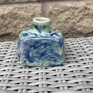 May include: A small, square-shaped ceramic bottle with a short, narrow neck. The surface features a mottled, hand-painted pattern of deep blue brushstrokes layered over a pale, light green base, creating a textured, marbled appearance.