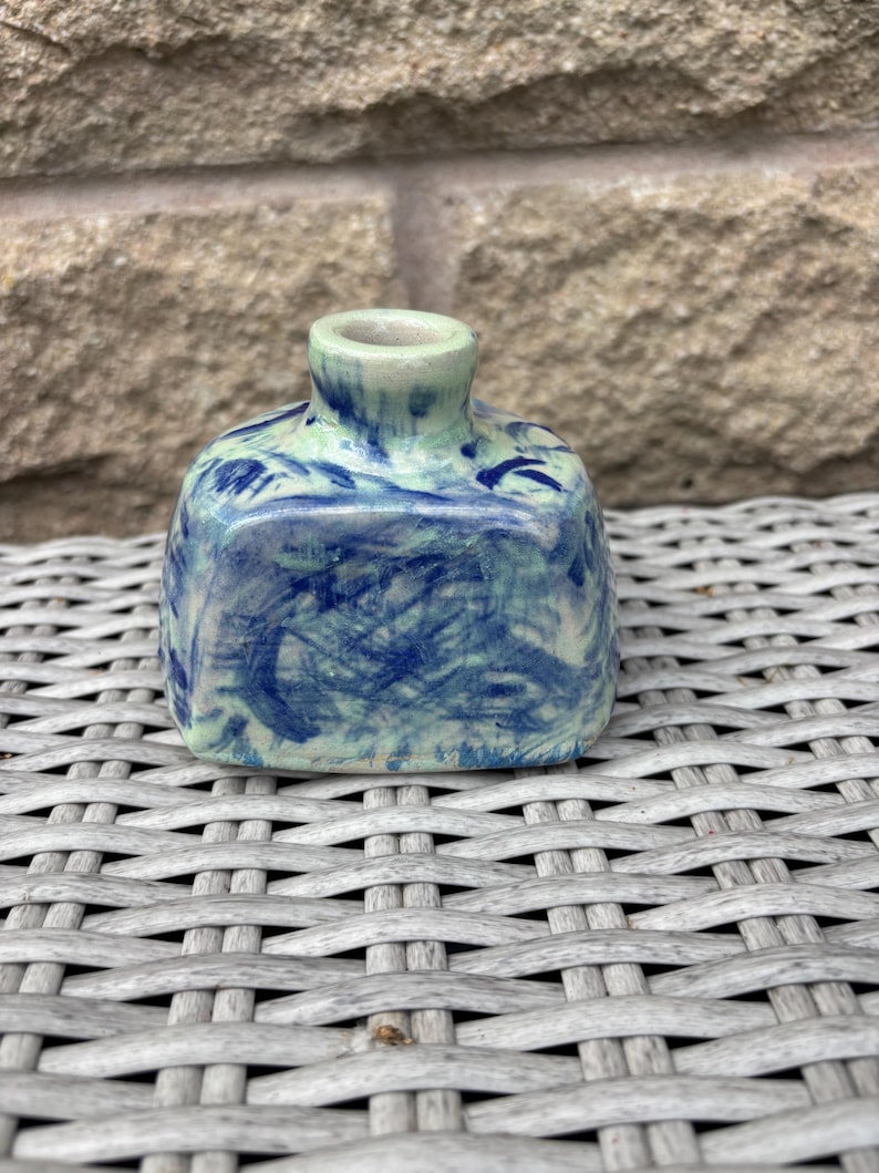 May include: A small, square-shaped ceramic bottle with a short, narrow neck. The surface features a mottled, hand-painted pattern of deep blue brushstrokes layered over a pale, light green base, creating a textured, marbled appearance.