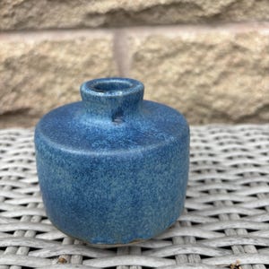 May include: A small, cylindrical ceramic vase with a mottled, deep blue glaze. The vase features a short, narrow neck and a flat, circular top surface that transitions into rounded sides, showcasing a textured, speckled finish throughout.