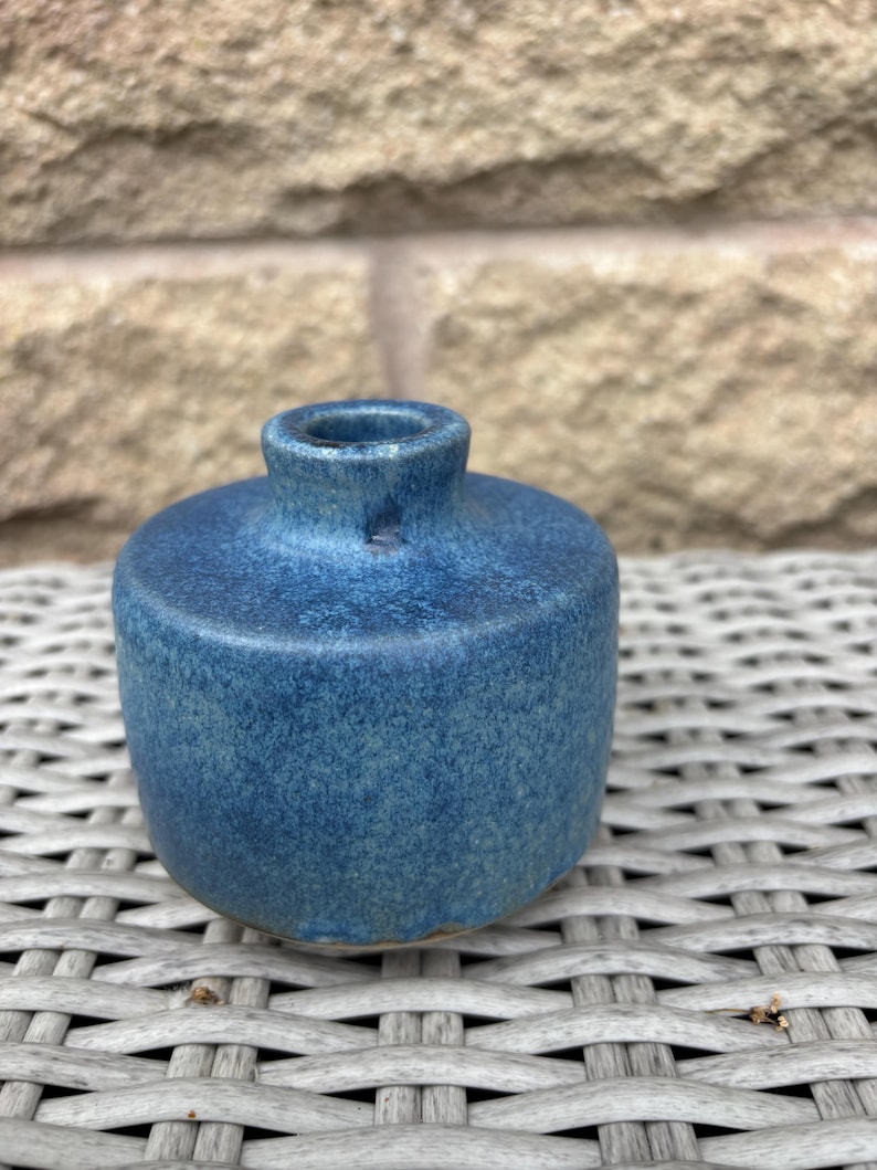 May include: A small, cylindrical ceramic vase with a mottled, deep blue glaze. The vase features a short, narrow neck and a flat, circular top surface that transitions into rounded sides, showcasing a textured, speckled finish throughout.