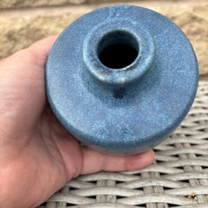 May include: A small, round ceramic vase with a textured, mottled blue glaze. The vase features a short, narrow neck and a circular opening at the top, showcasing a deep, dark interior. The surface has a matte finish with subtle variations in blue tones.
