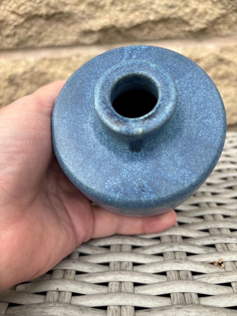 May include: A small, round ceramic vase with a textured, mottled blue glaze. The vase features a short, narrow neck and a circular opening at the top, showcasing a deep, dark interior. The surface has a matte finish with subtle variations in blue tones.