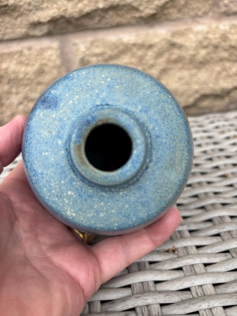 May include: A round ceramic vase with a speckled blue and light green glaze. The vase features a narrow, circular opening at the top, showcasing a textured finish with subtle variations in colour across the spherical body of the vessel.