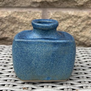 May include: A small, rectangular ceramic vase with a rounded top and a short, centred neck. The piece features a mottled, speckled blue glaze with subtle variations in tone, ranging from deep navy to lighter, dusty blue hues across the surface.