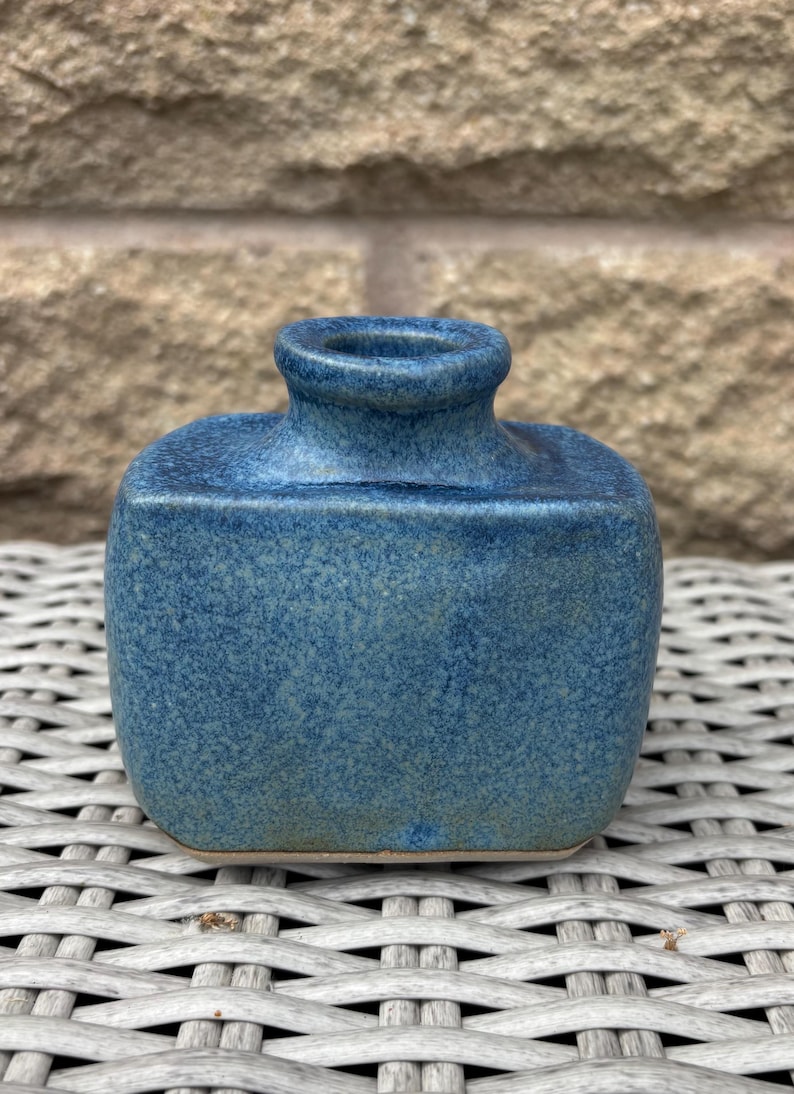 May include: A small, rectangular ceramic vase with a rounded top and a short, centred neck. The piece features a mottled, speckled blue glaze with subtle variations in tone, ranging from deep navy to lighter, dusty blue hues across the surface.