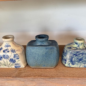 Puede incluir: Una colección de cinco pequeños jarrones de cerámica en varias formas y acabados en tonos azules. Dos jarrones son de color azul sólido con formas redondeadas o rectangulares, mientras que los otros tienen bases crema con patrones florales.
