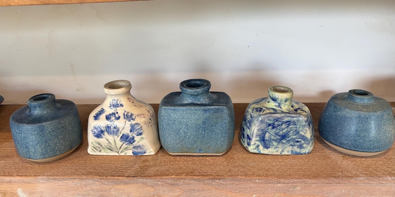 May include: A collection of five small ceramic bud vases in various shapes and blue-toned finishes. Two vases are solid blue with rounded or rectangular forms, while the others feature cream bases with blue floral or abstract brushstroke patterns.