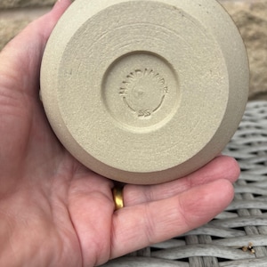 May include: A circular, light beige ceramic base with a matte, textured finish. The centre features a circular indentation containing the embossed text "HANDMADE" arched along the top and the letters "SS" positioned directly below the text.