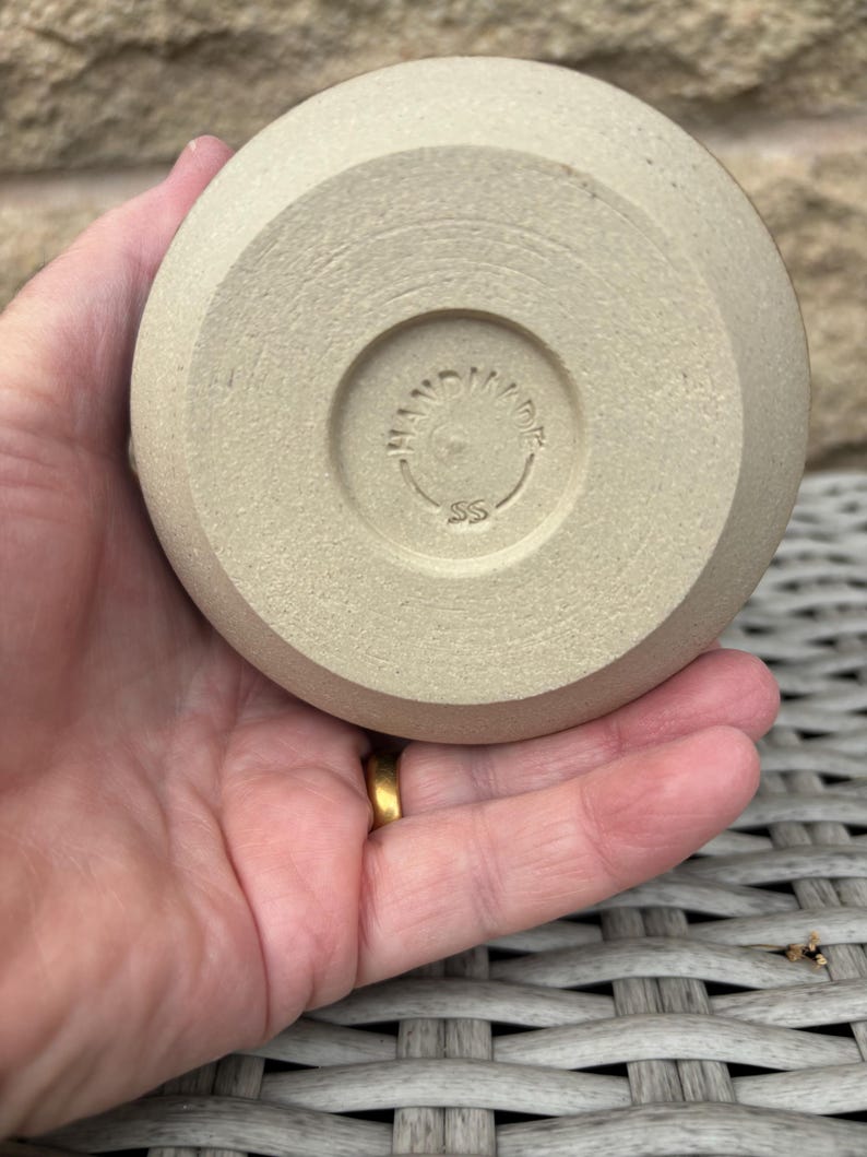 May include: A circular, light beige ceramic base with a matte, textured finish. The centre features a circular indentation containing the embossed text "HANDMADE" arched along the top and the letters "SS" positioned directly below the text.