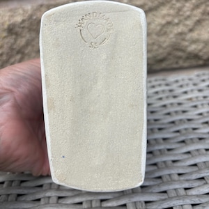 May include: A rectangular ceramic base with a matte, off-white finish. The surface features a circular stamp containing the text HANDMADE, a heart symbol in the centre, and the letters SS positioned below the heart, all impressed into the clay.
