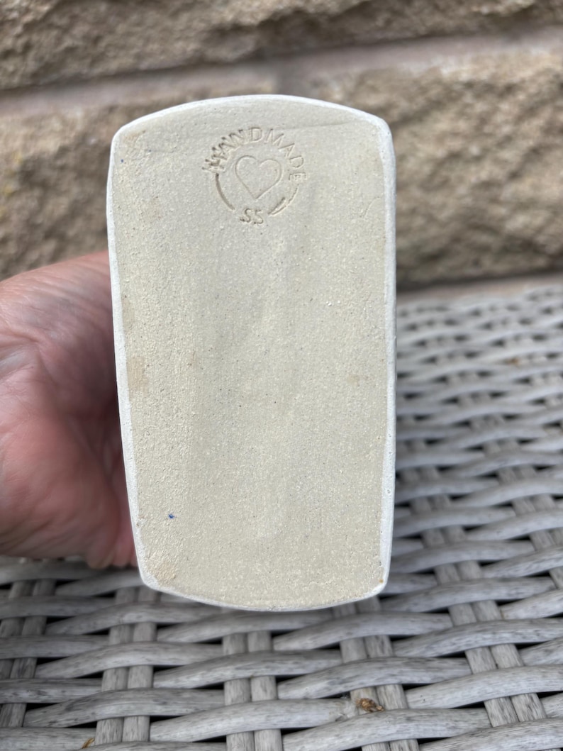 May include: A rectangular ceramic base with a matte, off-white finish. The surface features a circular stamp containing the text HANDMADE, a heart symbol in the centre, and the letters SS positioned below the heart, all impressed into the clay.