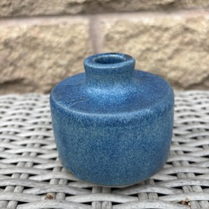 May include: A small, cylindrical ceramic vase with a speckled blue glaze. The vessel features a flat top surface, a short, narrow neck, and a circular opening. The finish displays variations in blue tones, ranging from deep navy to lighter azure.