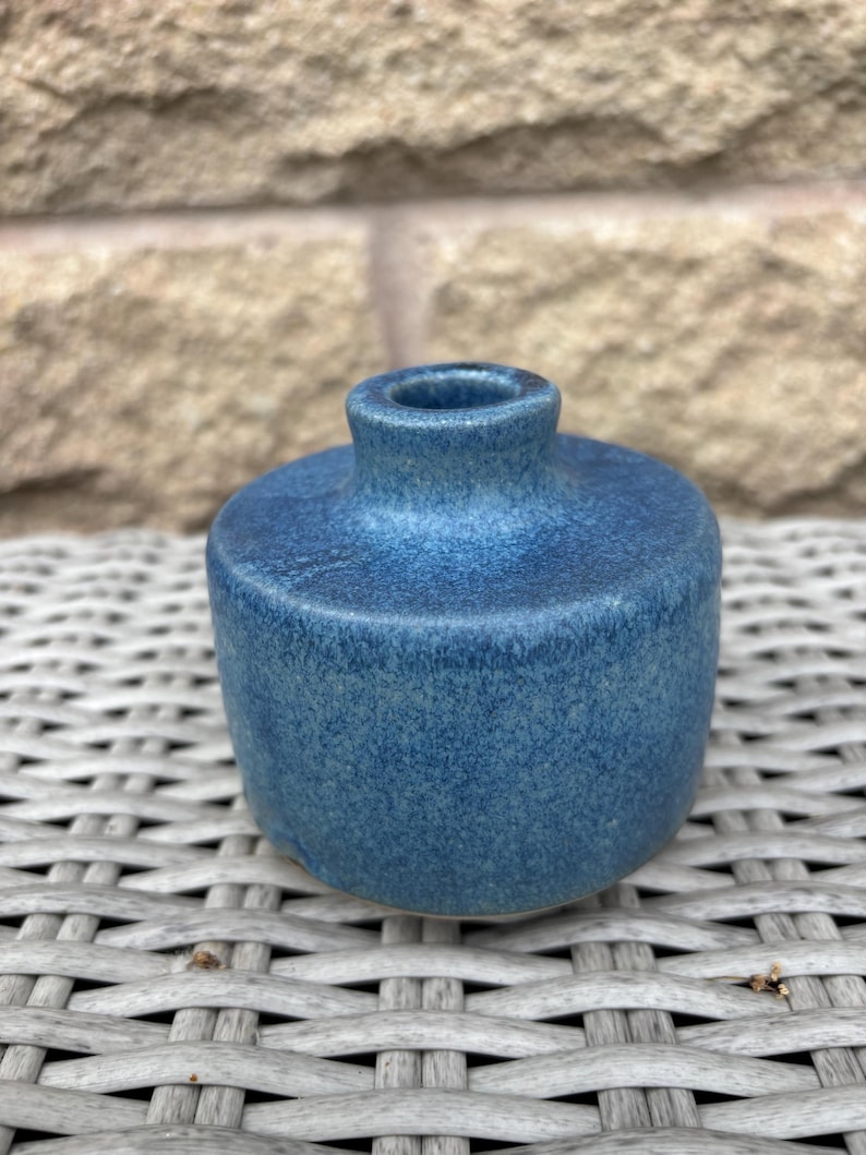 May include: A small, cylindrical ceramic vase with a speckled blue glaze. The vessel features a flat top surface, a short, narrow neck, and a circular opening. The finish displays variations in blue tones, ranging from deep navy to lighter azure.