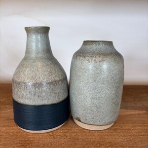 May include: Two ceramic vases. One is a tall, slender bottle shape with a dark blue band at the base. The other is a shorter, rounded vase. Both have a speckled grey finish and a neutral colour rim.