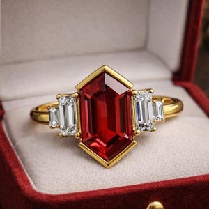 May include: A gold ring featuring a large, central, hexagonal-cut red gemstone flanked by two smaller, rectangular baguette-cut clear stones on each side. The ring is displayed inside a deep red velvet jewelry box with a light-colored interior lining.