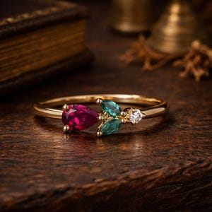 Puede incluir: Un anillo de oro con un rubí en forma de pera, dos gemas de talla marquesa de color verde azulado y un pequeño diamante. El anillo está engarzado en una fina banda de oro. Las gemas están dispuestas en un patrón floral.