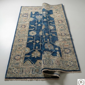 Pode incluir: Um tapete retangular enrolado, com um campo central azul e uma borda bege. O tapete apresenta padrões florais e geométricos em tons de azul, bege e cinza. As bordas têm franjas.