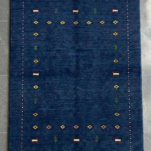 May include: A rectangular, navy blue rug with a textured surface. The rug features a border of small white dots and a repeating pattern of yellow diamonds, green trees, and red and white rectangles. The rug is designed for indoor use.